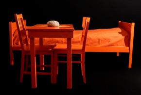 Ein Bett, ein Tisch mit drei Stühlen, alles in orange. auf dem Tisch liegt ein Laib Brot.