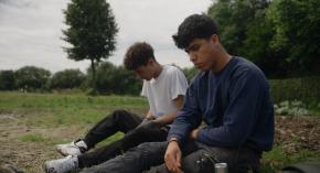 Die beiden jugendlichen Protagonisten des film sitzen auf einer Wiese und schauen zu Boden. sitzen auf einer 