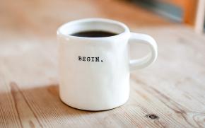 Eine weiße, mit schwarzem Kaffe gefüllte Tasse steht auf einem Holztisch. Auf der Tasse steht "Begin".